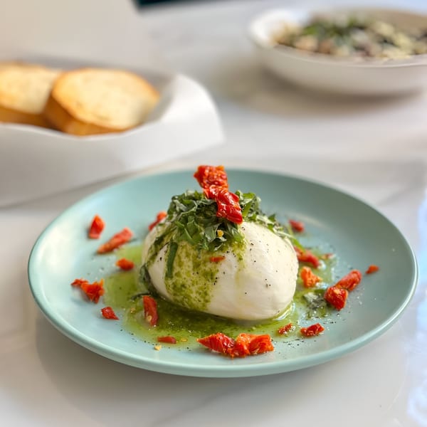 Burrata cheese with pesto, sun-dried tomatoes, and cracked pepper on a light blue plate.