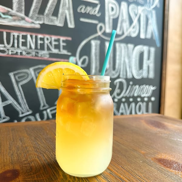 A refreshing iced drink with an orange slice and blue straw on a wooden table, with a chalkboard menu in the background.