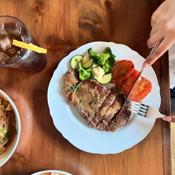 Overhead view of a person cutting a steak with roasted vegetables and iced tea on a wooden table.