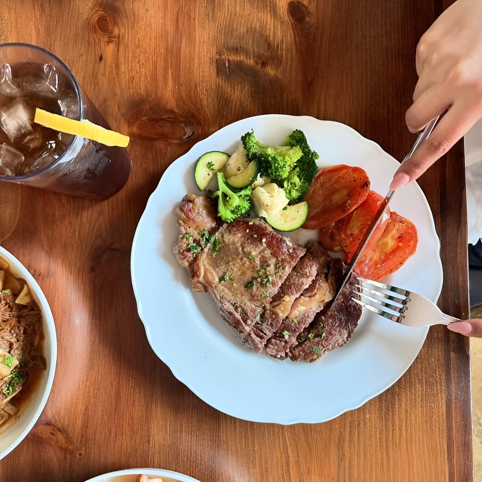 Overhead view of a person cutting a steak with roasted vegetables and iced tea on a wooden table.