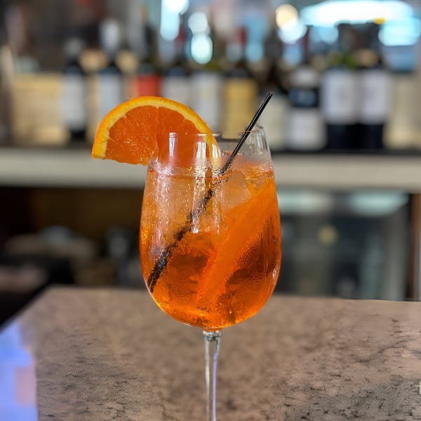 A vibrant orange Aperol Spritz cocktail with an orange slice and black straw on a bar.