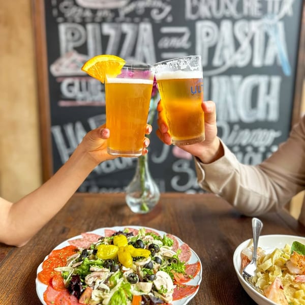 Two people toasting beers over a table with Petrini's Italian salad and pasta.