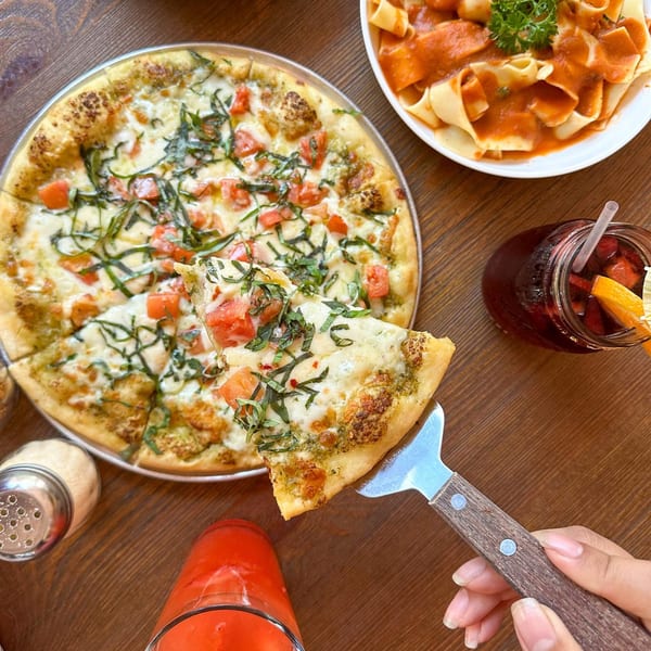 A hand serves a slice of Petrini's Italian pizza with tomatoes and basil, alongside pasta and drinks.